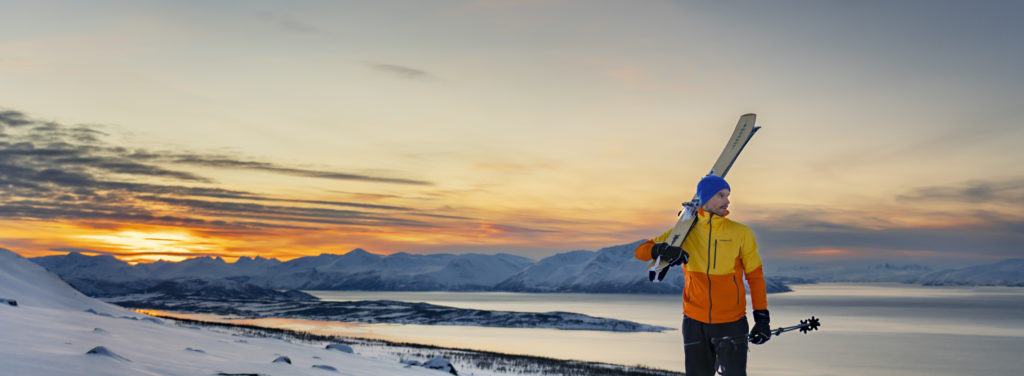 A skier on the mountain, with the view over the fjord, under a glowing winter sky. 